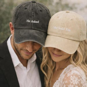 May include: A pair of baseball caps, one charcoal grey and one beige, with white embroidered text. The dark grey cap reads "He Asked," and the beige cap reads "She Said Yes." The caps are worn by a couple, suggesting a wedding or engagement theme.
