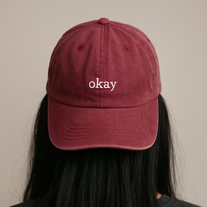 May include: A burgundy baseball cap with the word "okay" embroidered in white. The cap has a curved brim and a fabric strap closure. The hat is made of a soft, durable material.