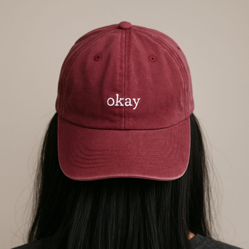 May include: A burgundy baseball cap with the word "okay" embroidered in white. The cap has a curved brim and a fabric strap closure. The hat is made of a soft, durable material.