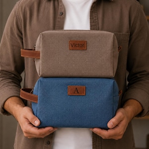Könnte beinhalten: Zwei rechteckige Kulturbeutel mit Seitengriffen, einer aus braunem und einer aus blauem Canvas. Jede Tasche hat einen braunen, lederartigen Aufnäher auf der Vorderseite. Die braune Tasche ist mit dem Namen Victor und die blaue mit dem Buchstaben A versehen.