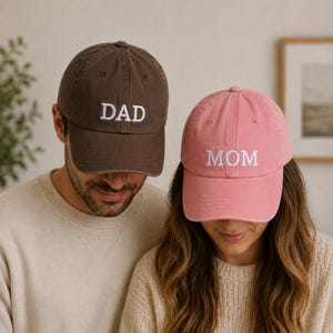 May include: Two baseball caps, one brown and one pink. The brown cap has the word "DAD" embroidered in white, and the pink cap has "MOM" embroidered in white. Both caps are worn by people.