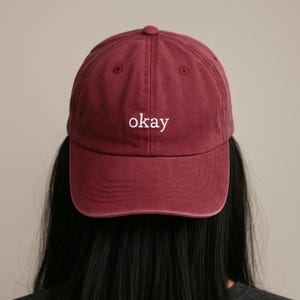 May include: A burgundy baseball cap with the word "okay" embroidered in white. The cap has a curved brim and a fabric strap closure. The hat is made of a soft, durable material.