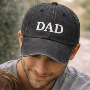May include: A dark gray baseball cap with the word "DAD" embroidered in white. The cap has a curved brim and a classic, casual style. The hat is made of a soft, worn material.