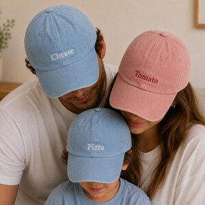 May include: Three baseball caps with embroidered text. One light blue cap features the word Cheese in white, a second light blue cap features the word Pizza in white, and a third dusty pink cap features the word Tomato in dark red thread.