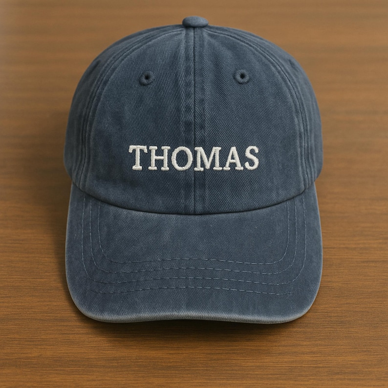 May include: A navy blue baseball cap with the name "THOMAS" embroidered in white. The cap has a curved peak and a button on top. The hat is made of a soft, textured fabric.
