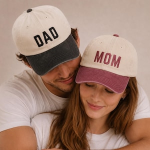 Könnte beinhalten: Zwei Baseballkappen. Eine ist cremefarben mit einem dunkelgrauen Schirm und dem Wort "DAD" in Schwarz gestickt. Die andere ist cremefarben mit einem bordeauxfarbenen Schirm und dem Wort "MOM" in Bordeauxrot gestickt.