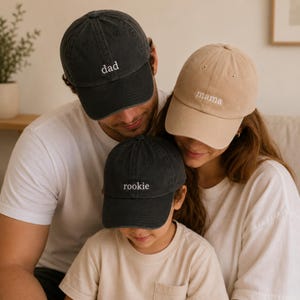 Peut inclure: Trois casquettes de baseball avec du texte brodé. Une casquette gris anthracite porte le mot dad, une casquette beige porte le mot mama, et une casquette gris anthracite porte le mot rookie. Chaque casquette a une visière courbée.