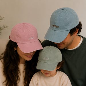 May include: Three baseball caps in muted colours. A pink cap with the word milk embroidered in white, a light blue cap with the word coffee embroidered in brown, and a sage green cap with the word latte embroidered in white. Each cap has a curved brim.