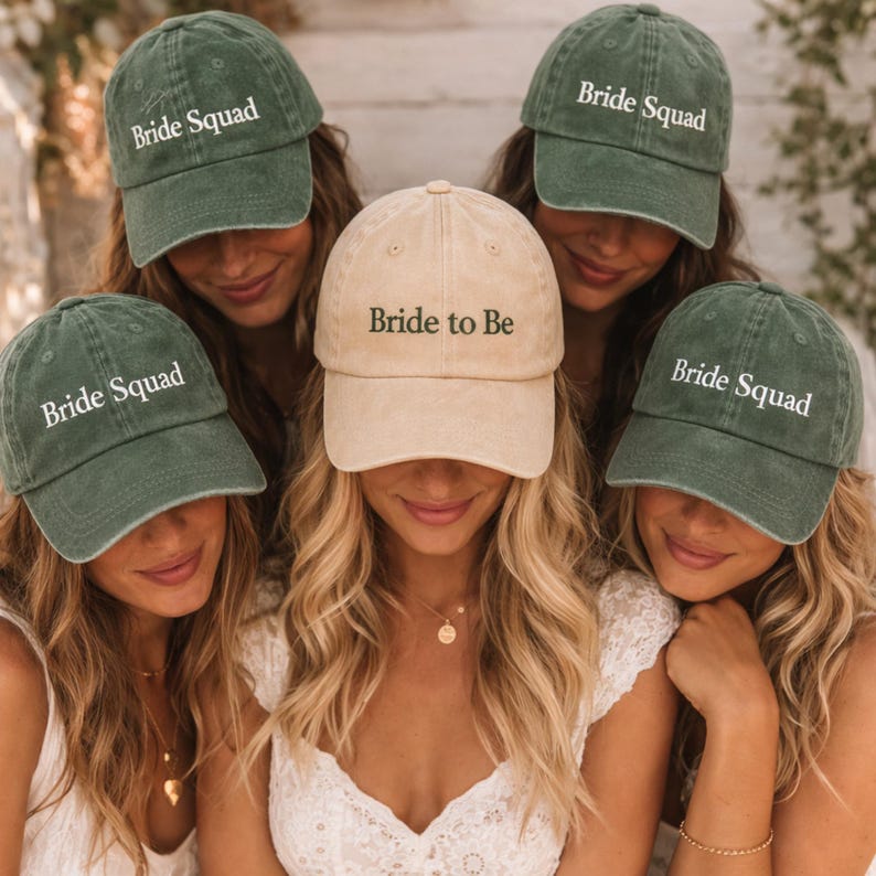 May include: A group of women wearing baseball caps. Four caps are olive green with "Bride Squad" in white text. One cap is tan with "Bride to Be" in white text. The women are wearing white tops and gold necklaces.