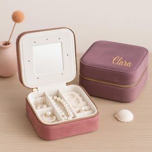 Op de afbeelding: Open, oudroze sieradendoos met een spiegel en vakken met parelsieraden. Een gesloten, bijpassende doos met "Clara" in goudkleurig schrift staat ernaast. Beide dozen hebben gouden ritsen.