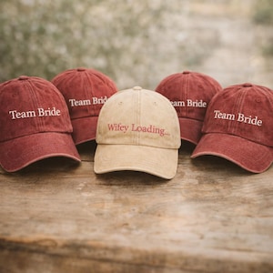 May include: Five baseball caps on a wooden surface. Four are maroon and say "Team Bride" in white text. The center cap is tan and reads "Wifey Loading..." in maroon text. The caps are a casual style.