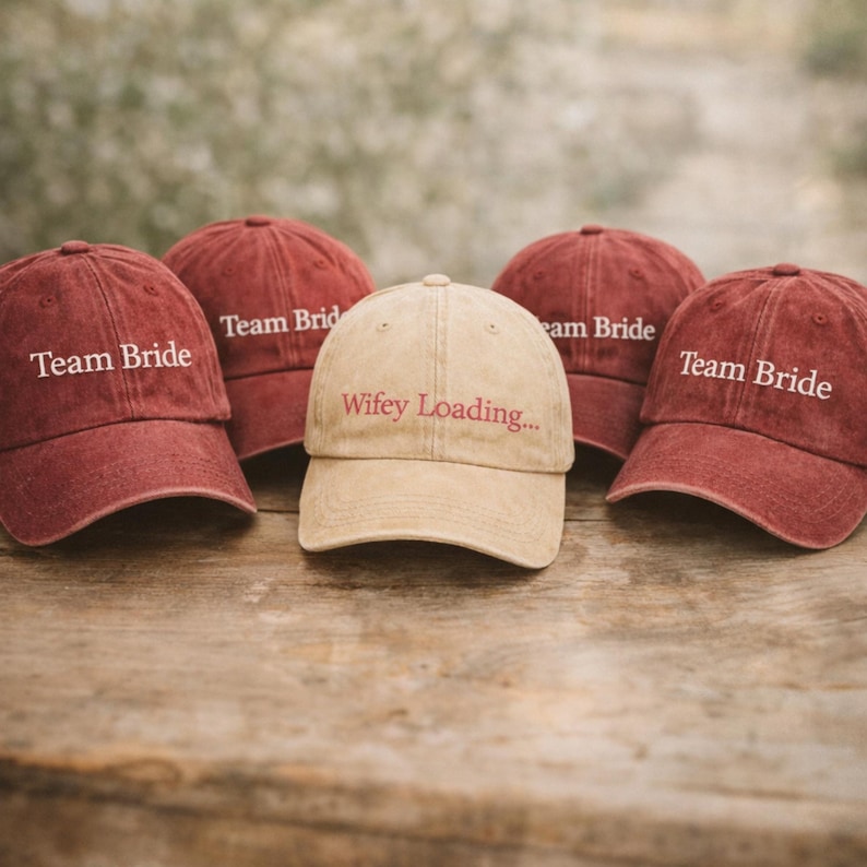 May include: Five baseball caps on a wooden surface. Four are maroon and say "Team Bride" in white text. The center cap is tan and reads "Wifey Loading..." in maroon text. The caps are a casual style.
