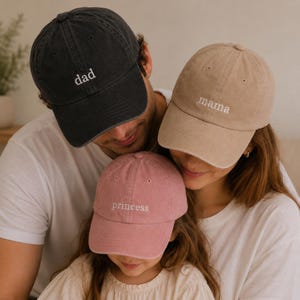 May include: Three baseball caps in charcoal, beige, and dusty pink. The charcoal cap features the white embroidered text "dad", the beige cap features the white embroidered text "mama", and the dusty pink cap features the white embroidered text "princess".