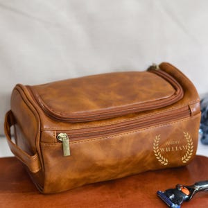 May include: A brown leather travel toiletry bag with a zipper closure and a gold handle. The bag has the name "Harry William" embossed on the front.