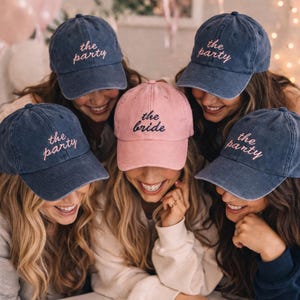 Könnte beinhalten: Eine Gruppe von Baseballkappen, eine rosa Kappe mit "the bride" und vier blaue Kappen mit "the party". Die Kappen sind mit kursivem Text bestickt. Das Bild zeigt eine Gruppe lächelnder Personen.