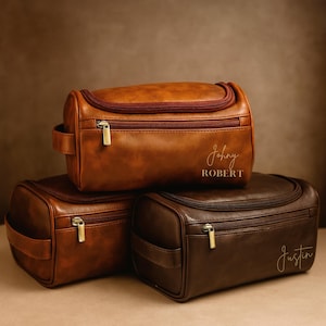 Op de afbeelding: Drie bruine leren toilettassen op elkaar gestapeld. De bovenste tas is lichtbruin, met de naam "Johny" en het woord "ROBERT" erop gedrukt. De middelste tas is middelbruin en de onderste tas is donkerbruin met de naam "Justin" erop gedrukt.