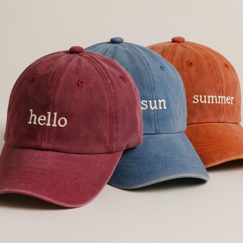 May include: Three baseball caps in red, blue, and orange. Each cap has a curved peak and a button on top. The red cap says "hello", the blue cap says "sun", and the orange cap says "summer".