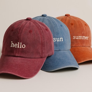 May include: Three baseball caps in shades of red, blue, and orange. Each cap has a curved brim and a small button on top. The red cap says "hello", the blue cap says "sun", and the orange cap says "summer".
