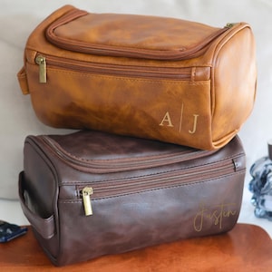 May include: Two brown leather travel toiletry bags with zipper closures. The top bag has the initials "A|J" embossed on the front. The bottom bag has the name "Justin" written in cursive on the front.