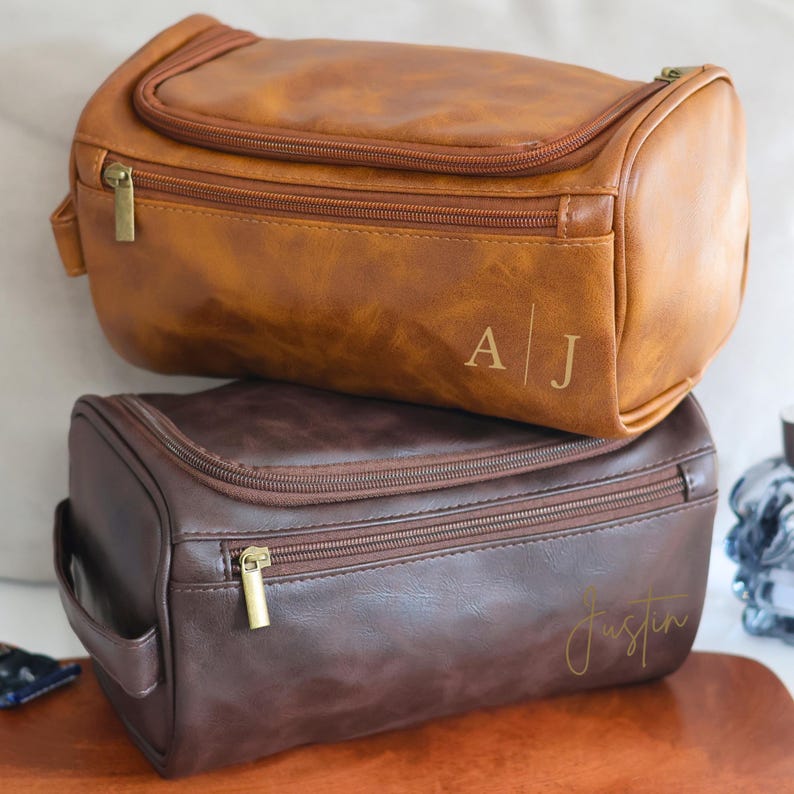 May include: Two brown leather travel toiletry bags with zipper closures. The top bag has the initials "A|J" embossed on the front. The bottom bag has the name "Justin" written in cursive on the front.