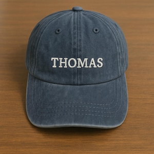 May include: A navy blue baseball cap with the name "THOMAS" embroidered in white block letters. The cap has a curved brim and a button on top. The hat is made of a soft, textured fabric.