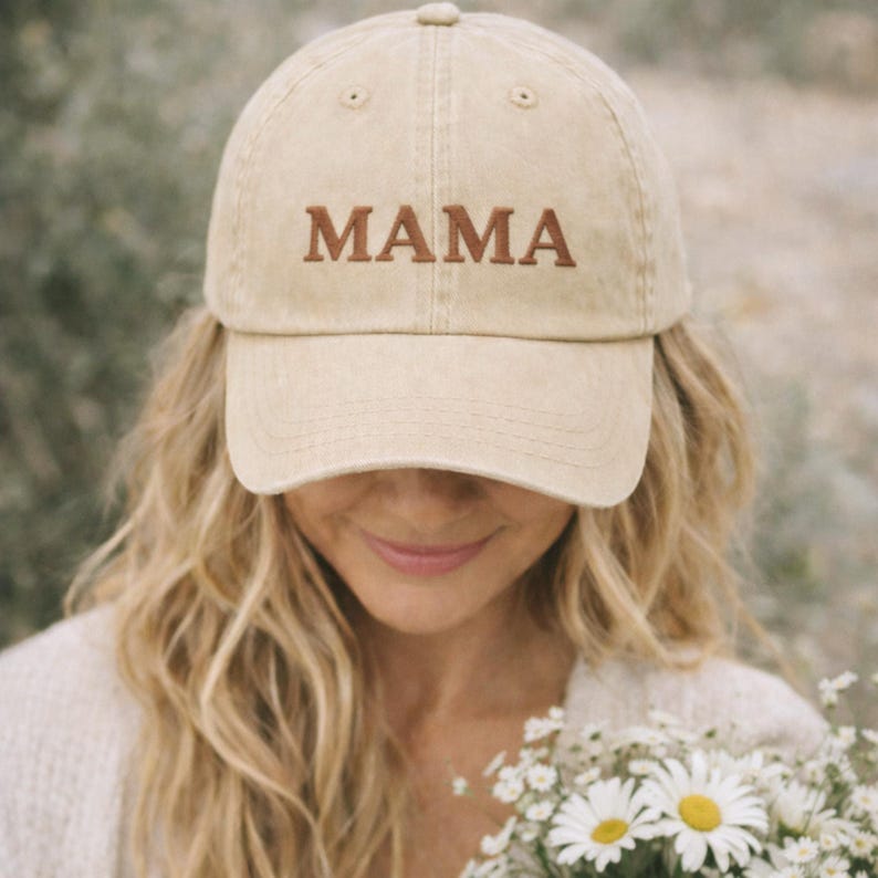 May include: A beige baseball cap embroidered with the word "MAMA" in brown. The cap is worn by a person holding a bouquet of white flowers with yellow centres.