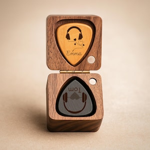 Könnte beinhalten: Eine offene Gitarrenplektrumbox aus Holz, die zwei Gitarrenplektren zeigt. Eines ist hellbraun mit Kopfhörer-Design und dem Namen "Emma". Das andere ist dunkelgrau mit Kopfhörer-Design und dem Namen "Tom".