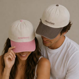 Peut inclure: Deux casquettes de baseball, une crème avec une visière rose et le mot "strawberry" brodé, l'autre crème avec une visière marron et le mot "chocolate" brodé. Les casquettes sont portées par deux personnes.
