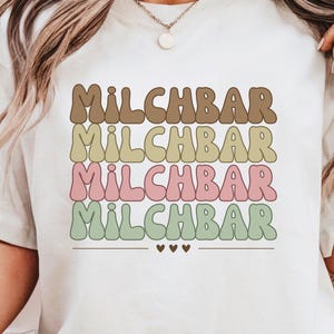 May include: White t-shirt with the text "MILCHBAR" repeated five times in a rainbow of colors, with brown, yellow, green, pink, and green. The text is in a retro font style.