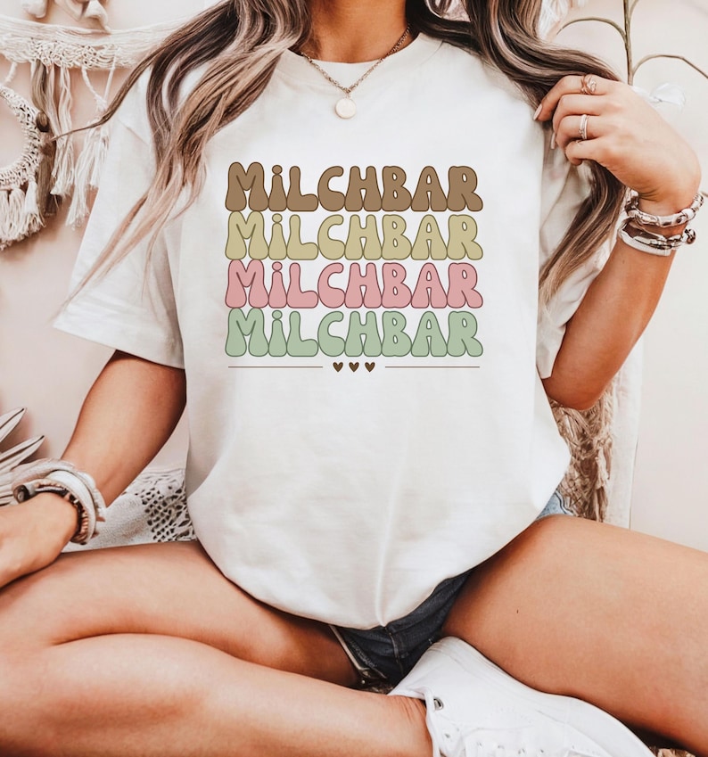 May include: White t-shirt with the text "MILCHBAR" repeated five times in a rainbow of colors, with brown, yellow, green, pink, and green. The text is in a retro font style.