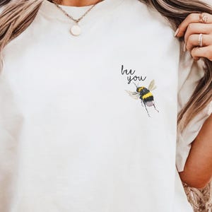 May include: White t-shirt with a bee graphic and the text "bee you" printed on the front.