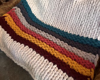 May include: A chunky knit blanket with a striped design. The blanket has a white base with stripes of teal, grey, yellow, red, and burgundy. The thick yarn gives a cosy, textured look, suitable for home decor.