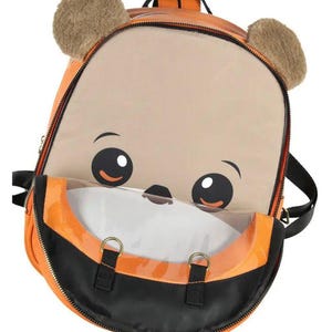 Puede incluir: Mochila naranja y negra con un dise&ntilde;o de cara de oso de dibujos animados. La mochila presenta un bolsillo frontal grande y transparente, orejas marrones y difusas y correas negras. La cremallera es negra y la mochila tiene herrajes met&aacute;licos dorados.