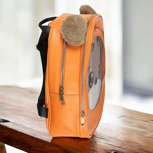 Puede incluir: Mochila naranja con un dise&ntilde;o de cara de perro de dibujos animados. La mochila tiene una ventana transparente que muestra la cara del perro, una oreja esponjosa y un peque&ntilde;o pomp&oacute;n. Cuenta con dos compartimentos con cremallera y correas negras. La mochila est&aacute; sobre una superficie de madera.