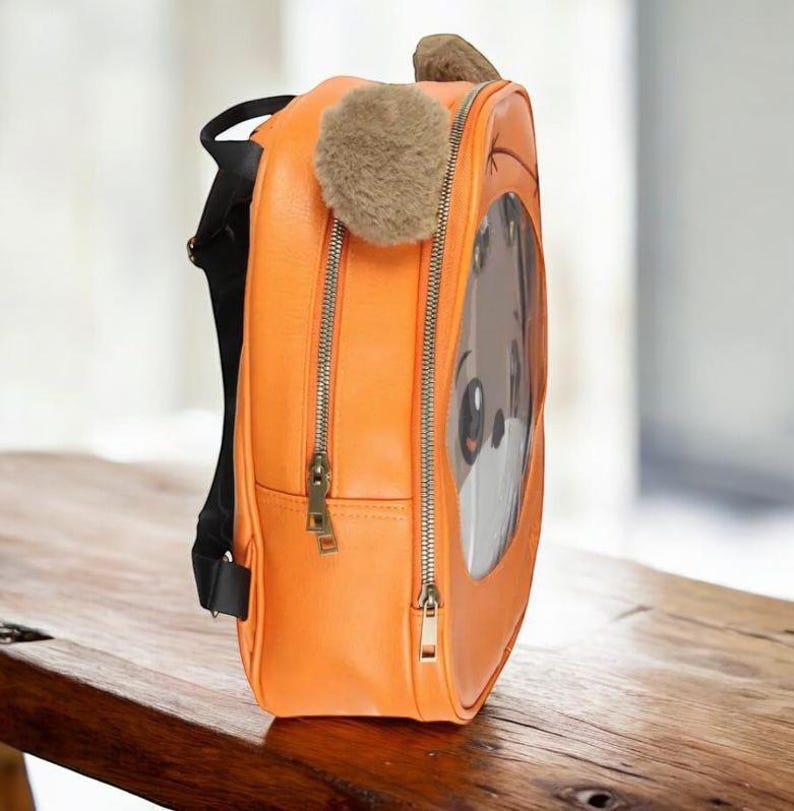 Puede incluir: Mochila naranja con un dise&ntilde;o de cara de perro de dibujos animados. La mochila tiene una ventana transparente que muestra la cara del perro, una oreja esponjosa y un peque&ntilde;o pomp&oacute;n. Cuenta con dos compartimentos con cremallera y correas negras. La mochila est&aacute; sobre una superficie de madera.