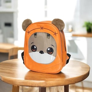 Puede incluir: Mochila naranja con un dise&ntilde;o de cara de oso de dibujos animados. La mochila presenta una ventana transparente que muestra la cara del oso, con ojos grandes y un hocico blanco. Dos acentos esponjosos marrones en forma de oreja se encuentran en la parte superior.