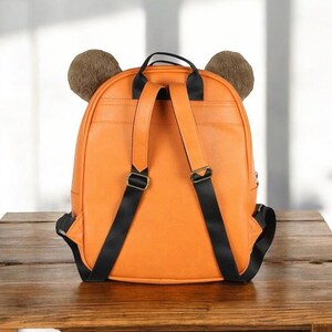 Puede incluir: Mochila naranja con correas negras y orejas de oso marrones. La mochila tiene un asa superior y correas ajustables para los hombros. La bolsa est&aacute; hecha de un material liso y se apoya sobre una superficie de madera.