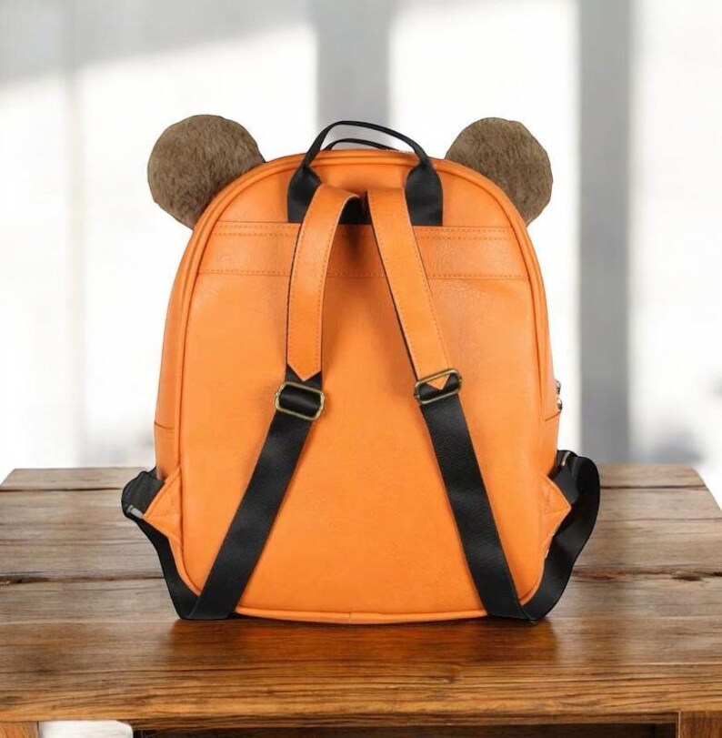 Puede incluir: Mochila naranja con correas negras y orejas de oso marrones. La mochila tiene un asa superior y correas ajustables para los hombros. La bolsa est&aacute; hecha de un material liso y se apoya sobre una superficie de madera.