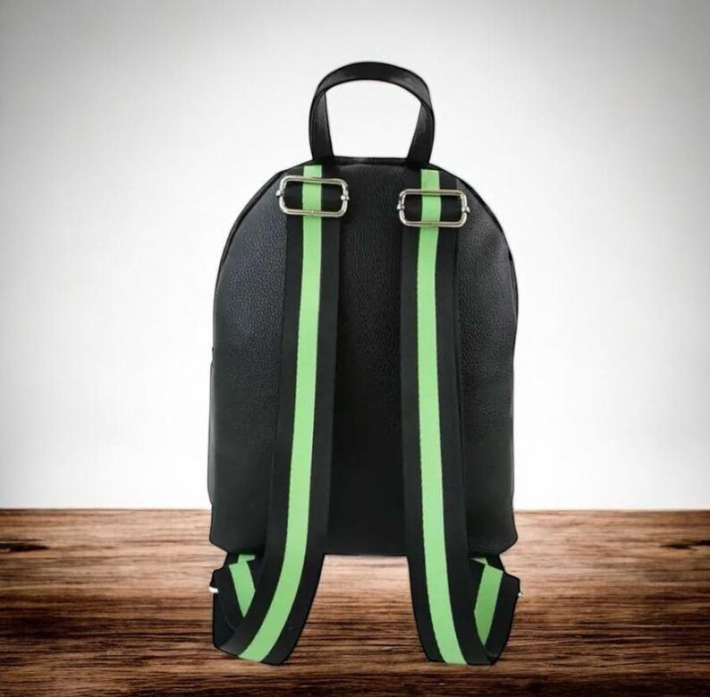 Puede incluir: Mochila negra con correas para los hombros a rayas verdes brillantes y negras. La mochila tiene un asa superior y hebillas de metal plateado. La bolsa est&aacute; hecha de un material texturizado.