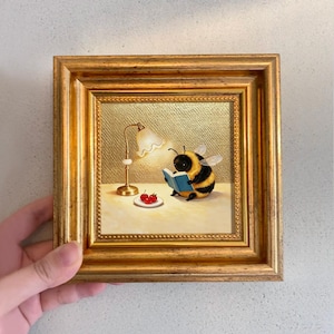 May include: A square painting in a gold frame depicting a fuzzy yellow and black bumblebee reading a small blue book under a brass desk lamp. Next to the bee is a white plate holding three red cherries on a light-colored surface with a gold background.