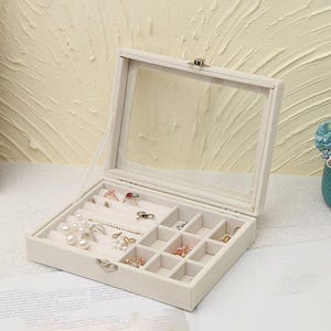 Könnte beinhalten: Eine beige Schmuckschatulle mit einem Deckel aus klarem Glas, geöffnet, um verschiedene Ringe, Halsketten und Ohrringe zu präsentieren. Die Schatulle ist in Fächer unterteilt und mit einem weichen, hellfarbigen Material ausgekleidet. Der Schmuck umfasst Perlen und goldfarbene Stücke.