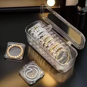 Peut inclure: Organisateur de bijoux en acrylique transparent avec plusieurs compartiments. L'organisateur contient des bracelets, des colliers et d'autres bijoux. Deux petits contenants carrés contiennent un bracelet de perles et un collier de perles. Sur une surface sombre.