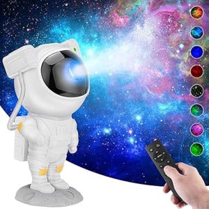 Puede incluir: Proyector de galaxia con forma de astronauta blanco, con un haz de luz azul. El proyector está sobre una base gris con forma de luna. Una mano sostiene un mando a distancia negro. El fondo muestra una galaxia colorida.