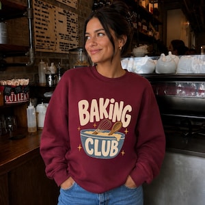 May include: A maroon crewneck sweatshirt featuring a graphic print of a blue mixing bowl with a whisk and wooden spoon. The text BAKING CLUB is printed in large cream-colored letters above and inside the bowl, accented by small yellow stars.