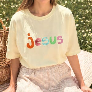 May include: A light yellow t-shirt with the word "JESUS" embroidered in colorful, patterned letters. The shirt has short sleeves and a crew neck. The image is taken outdoors, with a basket and flowers in the background.