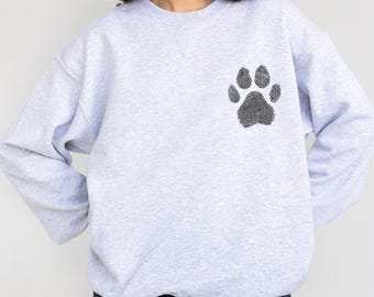 May include: A light gray sweatshirt featuring a black paw print design on the upper left chest. The paw print is made up of fingerprint patterns. The sweatshirt has long sleeves and a crew neck.