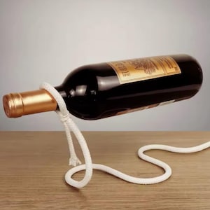 Puede incluir: Un soporte para botellas de vino de cuerda blanca con una botella de vino tinto suspendida en el aire. La botella tiene una etiqueta de papel de aluminio dorado con la palabra "Vielle" impresa en ella.