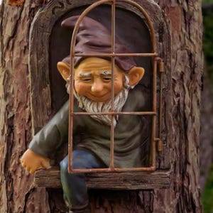 Puede incluir: Un gnomo con un sombrero marrón y barba asoma por una ventana en un árbol. La ventana es de madera y tiene una reja de metal.