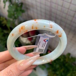 May include: A smooth, pale green jade bangle bracelet featuring scattered orange-red inclusions throughout the stone. A clear plastic tag with a QR code and the text 'NATURAL A CUI JADE' is attached to the circular band with a plastic fastener.