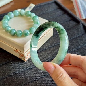 Op de afbeelding: Een gladde, gepolijste jade armband met donkergroene en doorschijnend lichtgroene marmerpatronen, vastgehouden naast een bijpassende jade kralenarmband die op een klein, vierkant, crèmekleurig doosje met bloemaccenten ligt.